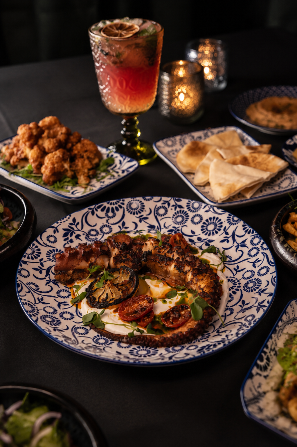 A candlelit table spread of charred octopus, mezze plates and a cocktail