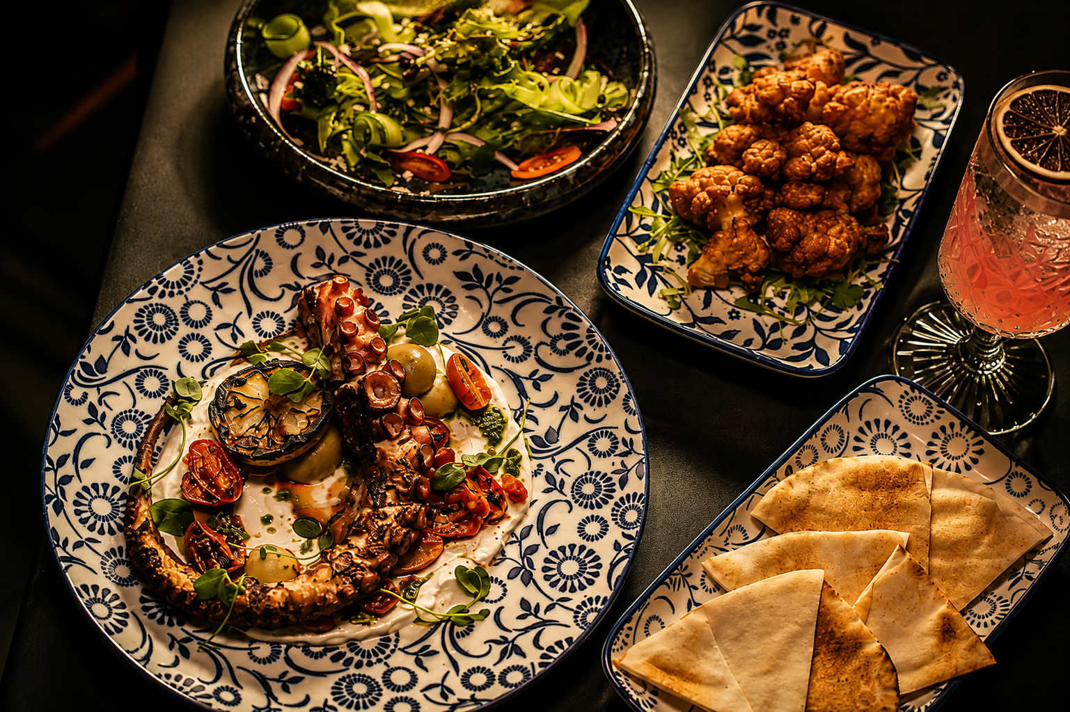 Table spread of grilled octopus, salad, crispy cauliflower, pita and a cocktail